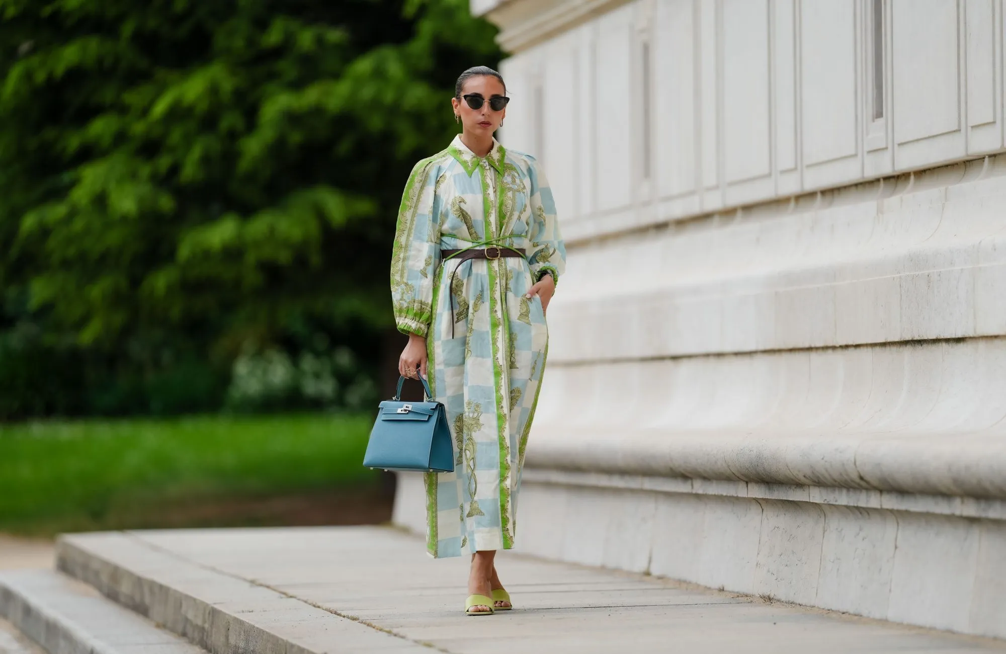 PARIS, FRANCE - MAY 12: Gabriella Berdugo wears sunglasses, earrings, a white blue and green checked long dress with long sleeves and floral print, a brown leather belt, a blue Kelly Hermes bag, pale pastel yellow mules / shoes with heart-shaped heels from Kat Maconie, during a street style fashion photo session, on May 12, 2024 in Paris, France. (Photo by Edward Berthelot/Getty Images)