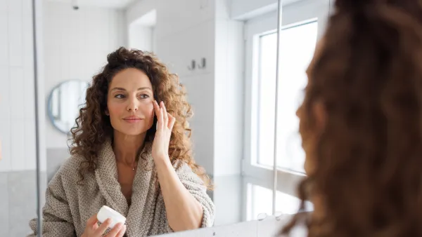 A mid adult woman with curly hair wearing a cozy bathrobe applies face cream to her face in a bright, modern bathroom with natural daylight streaming through the window.