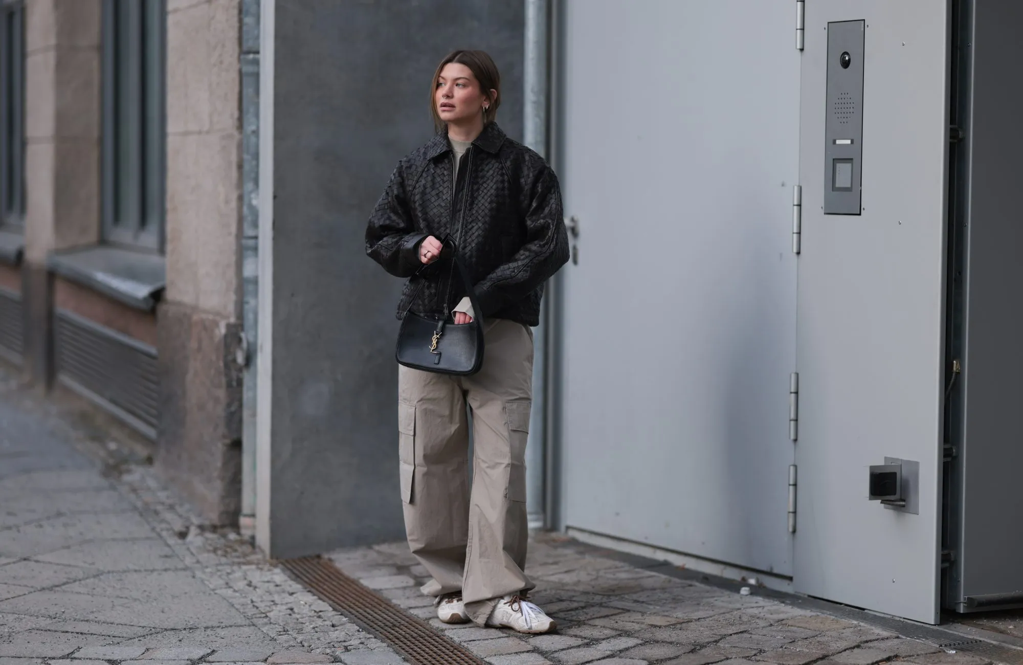 BERLIN, GERMANY - FEBRUARY 19: Sophia Geiss seen wearing a black woven leather jacket by Second Female layered over a minimalist H&M Studio look featuring a light beige high-neck top and oversized cargo trousers in a matching neutral tone. She pairs the outfit with Miu Miu x New Balance sneakers in white with brown detailing. Accessories include a structured black YSL black shoulder bag with gold hardware, Mango sunglasses held in her hand, and subtle silver hoop earrings. Her hair is styled in a loose low updo on February 19, 2026 in Berlin, Germany. (Photo by Jeremy Moeller/Getty Images)