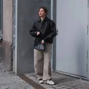 BERLIN, GERMANY - FEBRUARY 19: Sophia Geiss seen wearing a black woven leather jacket by Second Female layered over a minimalist H&M Studio look featuring a light beige high-neck top and oversized cargo trousers in a matching neutral tone. She pairs the outfit with Miu Miu x New Balance sneakers in white with brown detailing. Accessories include a structured black YSL black shoulder bag with gold hardware, Mango sunglasses held in her hand, and subtle silver hoop earrings. Her hair is styled in a loose low updo on February 19, 2026 in Berlin, Germany. (Photo by Jeremy Moeller/Getty Images)