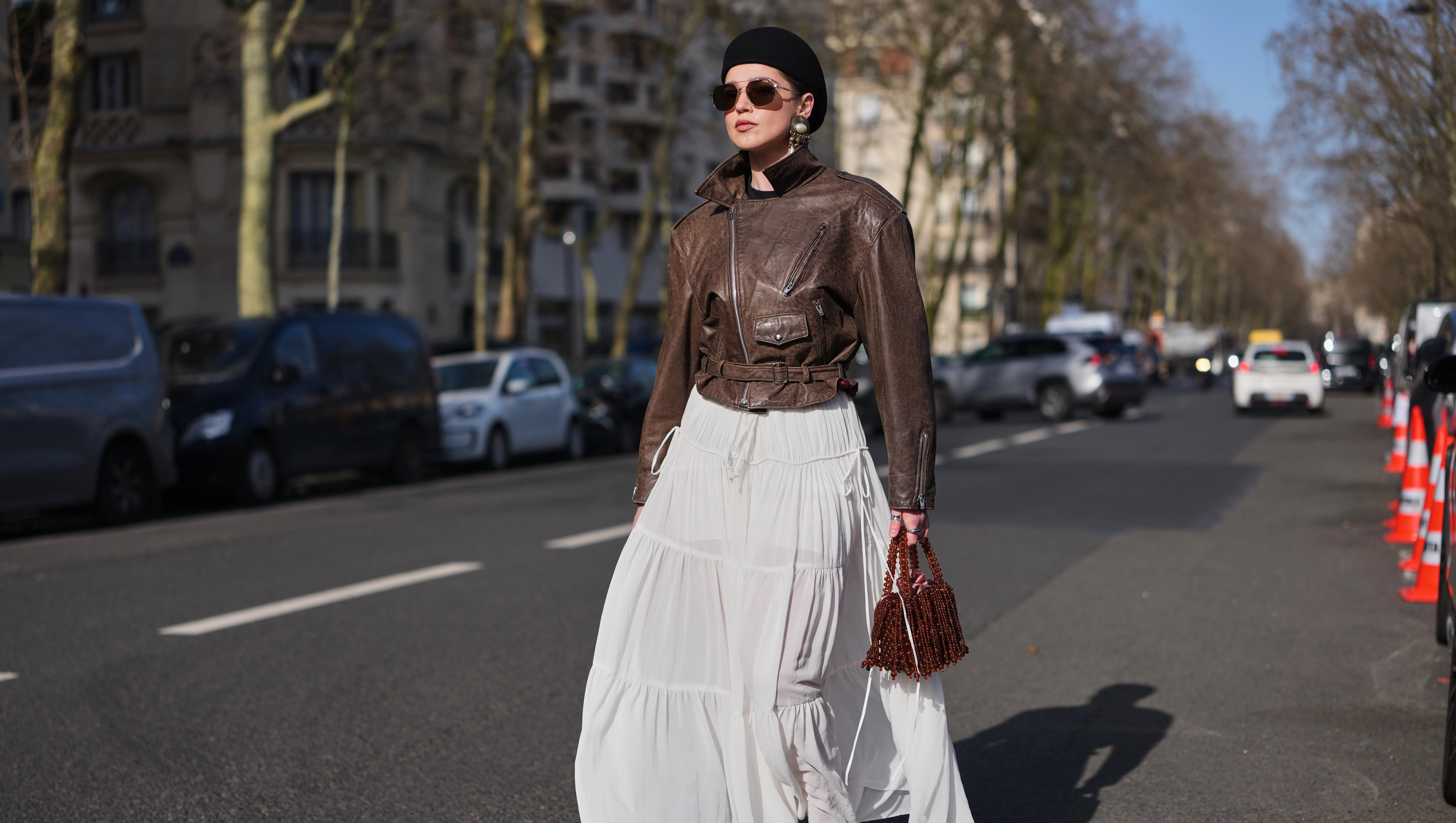 PARIS, FRANCE - MARCH 05: A guest wears a black felt pillbox hat, brown hair, brown tinted aviator sunglasses, a dark brown leather belted biker zipper jacket, a white chiffon tiered long skirt, a dark brown fringed beaded / pearls handbag, dark brown leather platform soles / high heels shoes, outside Chloe, during Paris Fashion Week - Womenswear Fall/Winter 2026/2027, on March 05, 2026 in Paris, France (Photo by Edward Berthelot/Getty Images)