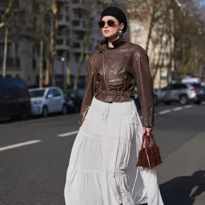 PARIS, FRANCE - MARCH 05: A guest wears a black felt pillbox hat, brown hair, brown tinted aviator sunglasses, a dark brown leather belted biker zipper jacket, a white chiffon tiered long skirt, a dark brown fringed beaded / pearls handbag, dark brown leather platform soles / high heels shoes, outside Chloe, during Paris Fashion Week - Womenswear Fall/Winter 2026/2027, on March 05, 2026 in Paris, France (Photo by Edward Berthelot/Getty Images)