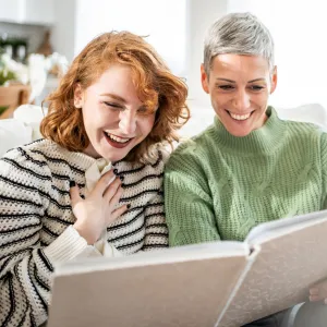 Happy mother and teenage daughter laughing while looking at a photo album, enjoying precious family memories together on a comfortable sofa in their living room