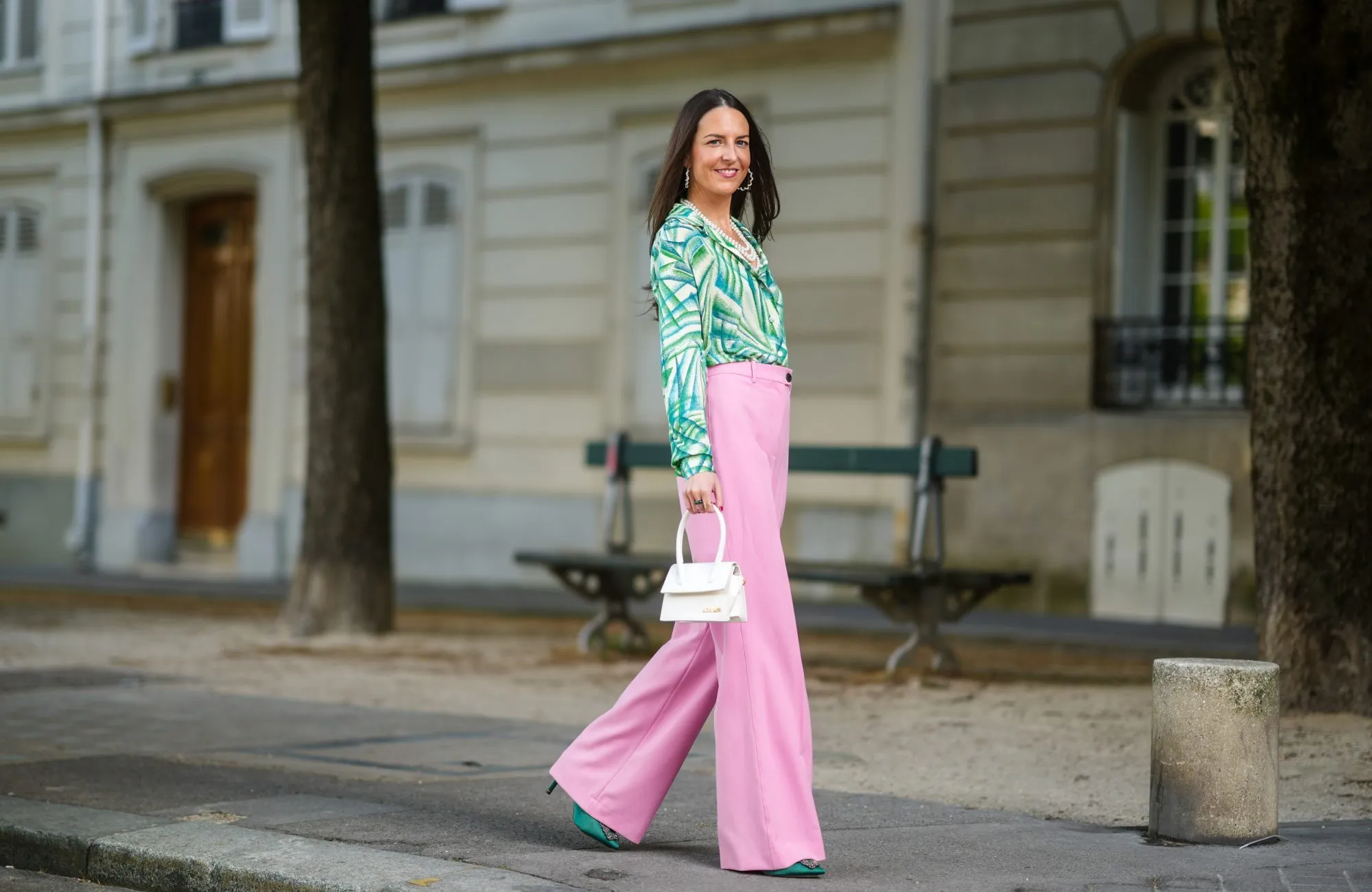 Alba Garavito Torre wears gold and pearls earrings, a white pearls necklace, a white with green tree leaf pattern shirt, pale pink flared suit pants, gold rings, a green ring, a pink ring, a gold Love Cartier bracelet, gold bracelets, a white matter leather Chiquito Medium Jacquemus handbag, dark green satin Manolo Blahnik pumps shoes, on May 23, 2021 in Paris, France.