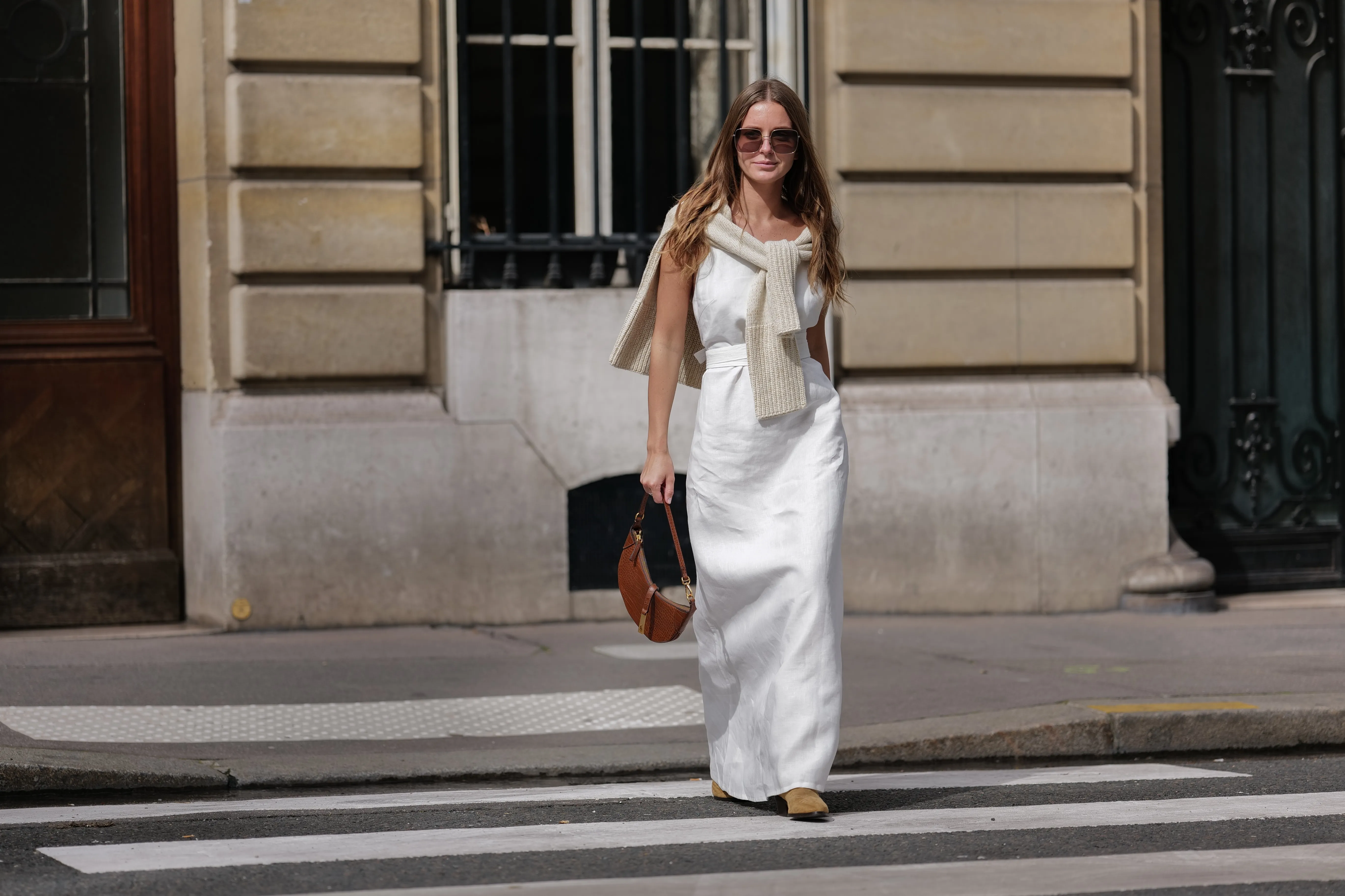Diane Batoukina wears dark brown large squared sunglasses from Dior, a white square-neck / linen belted long dress, a white latte ribbed wool pullover knot at the shoulder, a camel brown shiny leather crocodile print pattern handbag from Ralph Lauren, brown suede block heels / pointed ankle boots , during a street style fashion photo session, on April 28, 2023 in Paris, France.