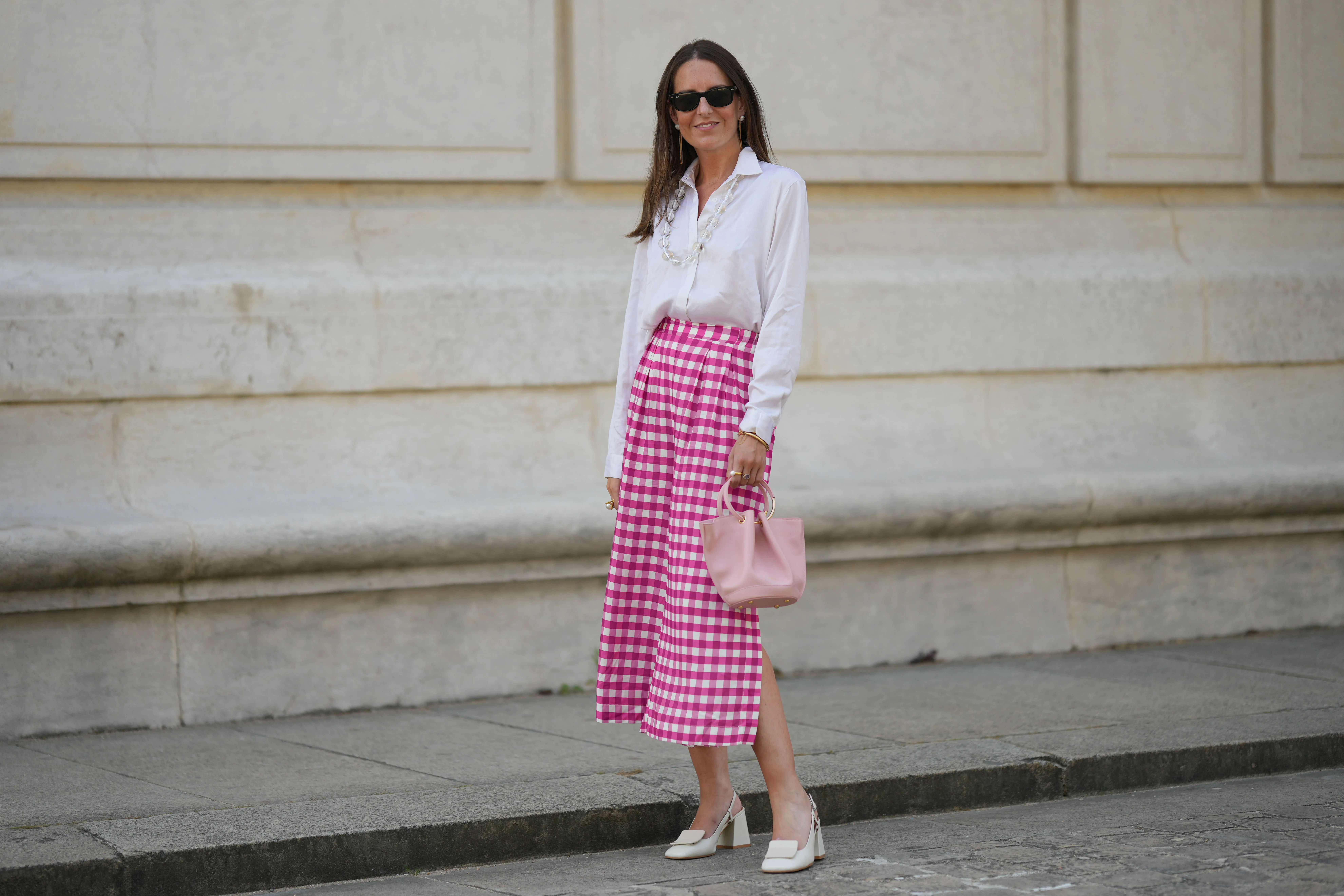 Alba Garavito Torre wears black sunglasses from Ray Ban, white pearl and gold chain pendant earrings, a transparent large pearls long necklace, a white shirt, pink and white checkered print pattern midi pants from Frambu&atilde;, gold bracelets, a pale pink matte leather handbag from Saonara Brand, white latte matte shiny leather block heels mules from Jonak, during a street style fashion photo session, on June 06, 2023 in Paris, France.