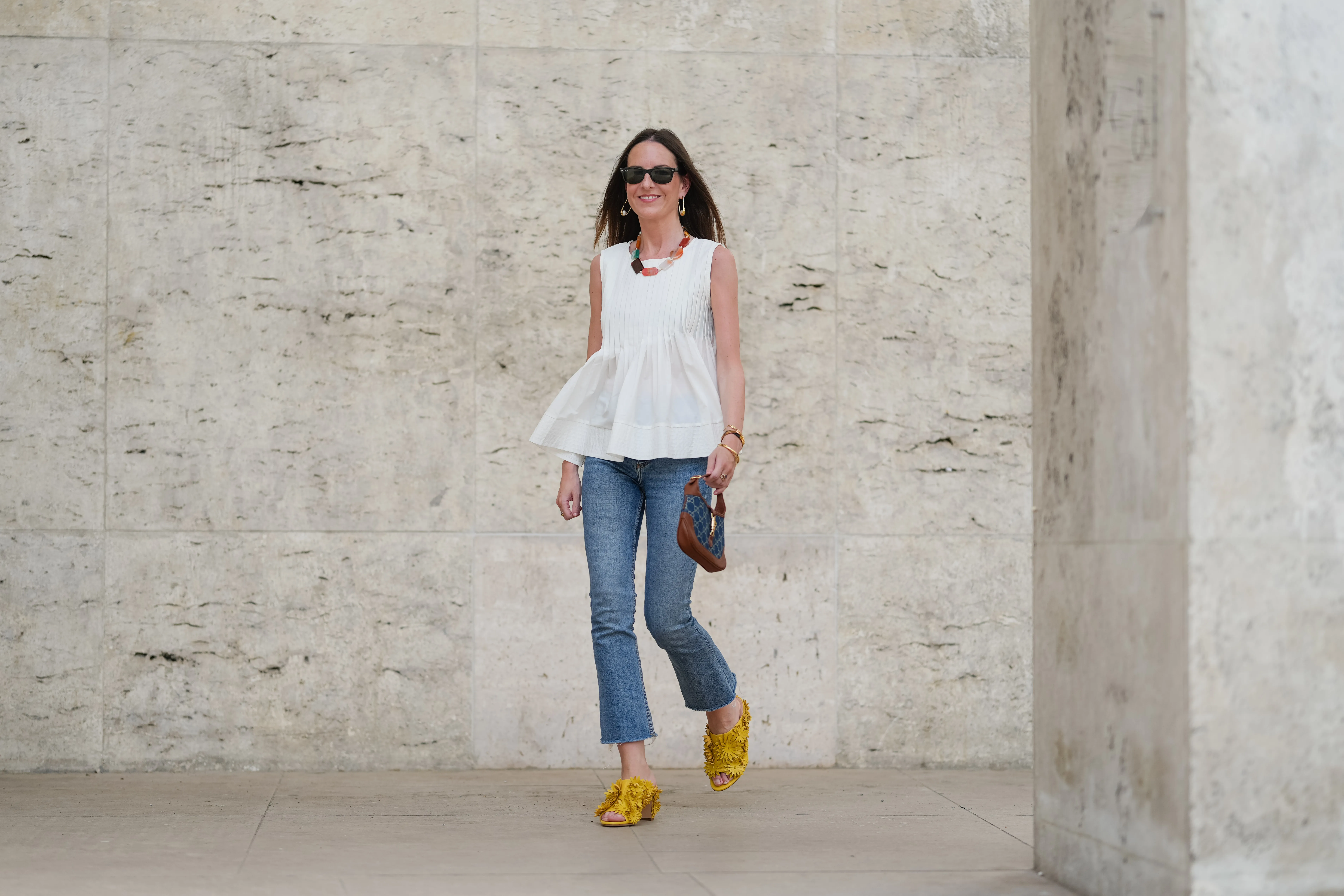 Alba Garavito Torre wears black sunglasses, long straight brown hair with a center part, a white pleated peplum sleeveless blouse by Commense, and blue high-rise raw-hem denim jeans. A yellow clog sandal with floral detailing by AGL is worn, paired with a jean denim and brown mini Jackie handbag by Gucci. Accessories include a chunky necklace with colorful natural stones by Sempiterno Paris and gold jewelry. Full body shot, during a street style fashion photo session, on May 02, 2025 in Paris, France.
