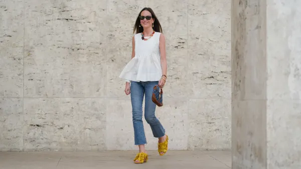 Alba Garavito Torre wears black sunglasses, long straight brown hair with a center part, a white pleated peplum sleeveless blouse by Commense, and blue high-rise raw-hem denim jeans. A yellow clog sandal with floral detailing by AGL is worn, paired with a jean denim and brown mini Jackie handbag by Gucci. Accessories include a chunky necklace with colorful natural stones by Sempiterno Paris and gold jewelry. Full body shot, during a street style fashion photo session, on May 02, 2025 in Paris, France.