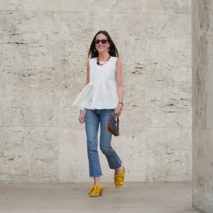 Alba Garavito Torre wears black sunglasses, long straight brown hair with a center part, a white pleated peplum sleeveless blouse by Commense, and blue high-rise raw-hem denim jeans. A yellow clog sandal with floral detailing by AGL is worn, paired with a jean denim and brown mini Jackie handbag by Gucci. Accessories include a chunky necklace with colorful natural stones by Sempiterno Paris and gold jewelry. Full body shot, during a street style fashion photo session, on May 02, 2025 in Paris, France.