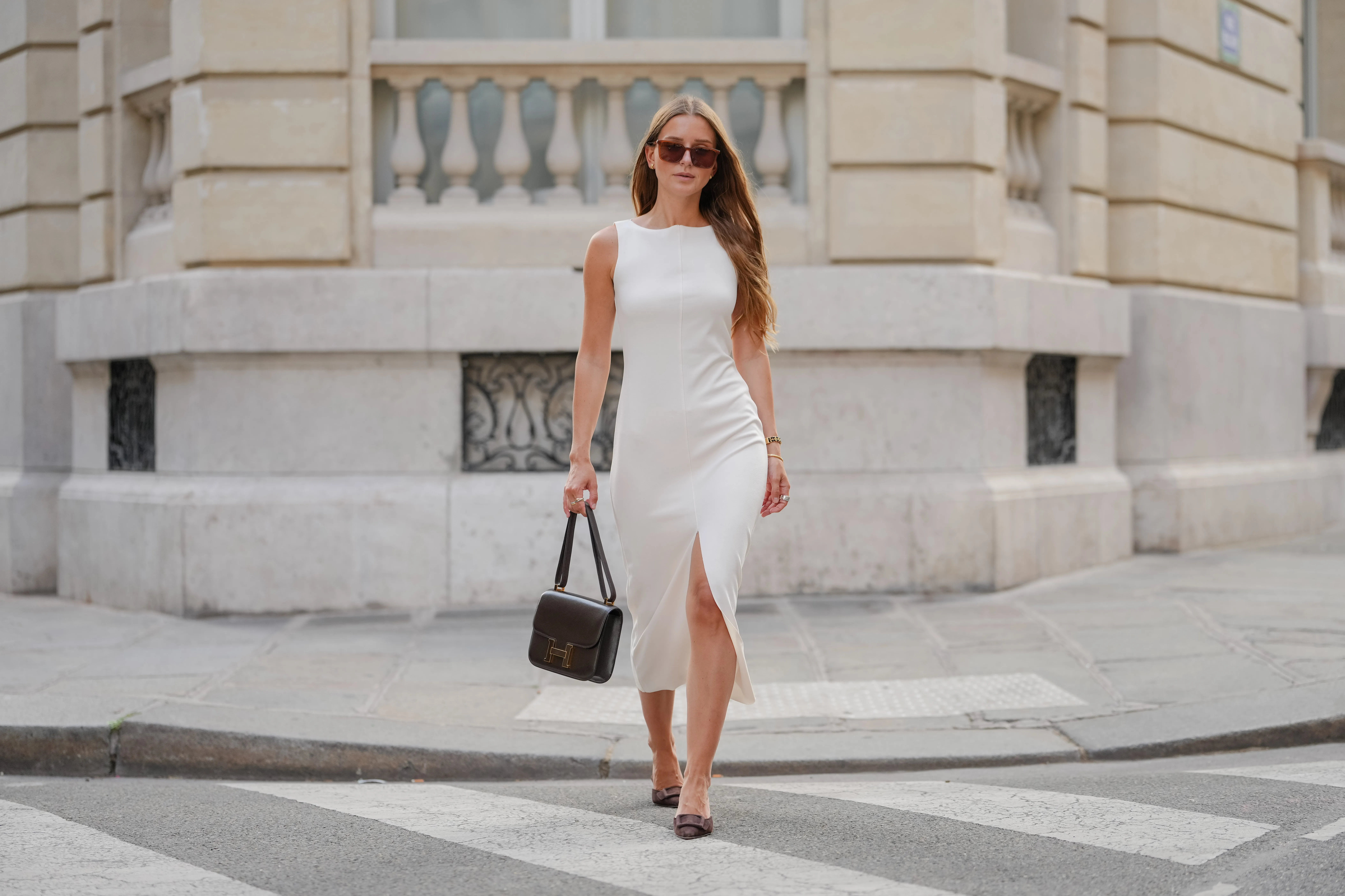 Diane Batoukina wears a sleeveless, fitted white dress with a high neckline and a midi length. Long, loose hair is styled with a center part. Sunglasses feature a brown tint and a rectangular shape. A black leather handbag from Hermes with a structured design and a prominent gold "H" detail is carried. On the wrist, a delicate gold bracelet is visible. Footwear consists of pointed-toe shoes in a dark color, during a street style fashion photo session, on June 13, 2025 in Paris, France