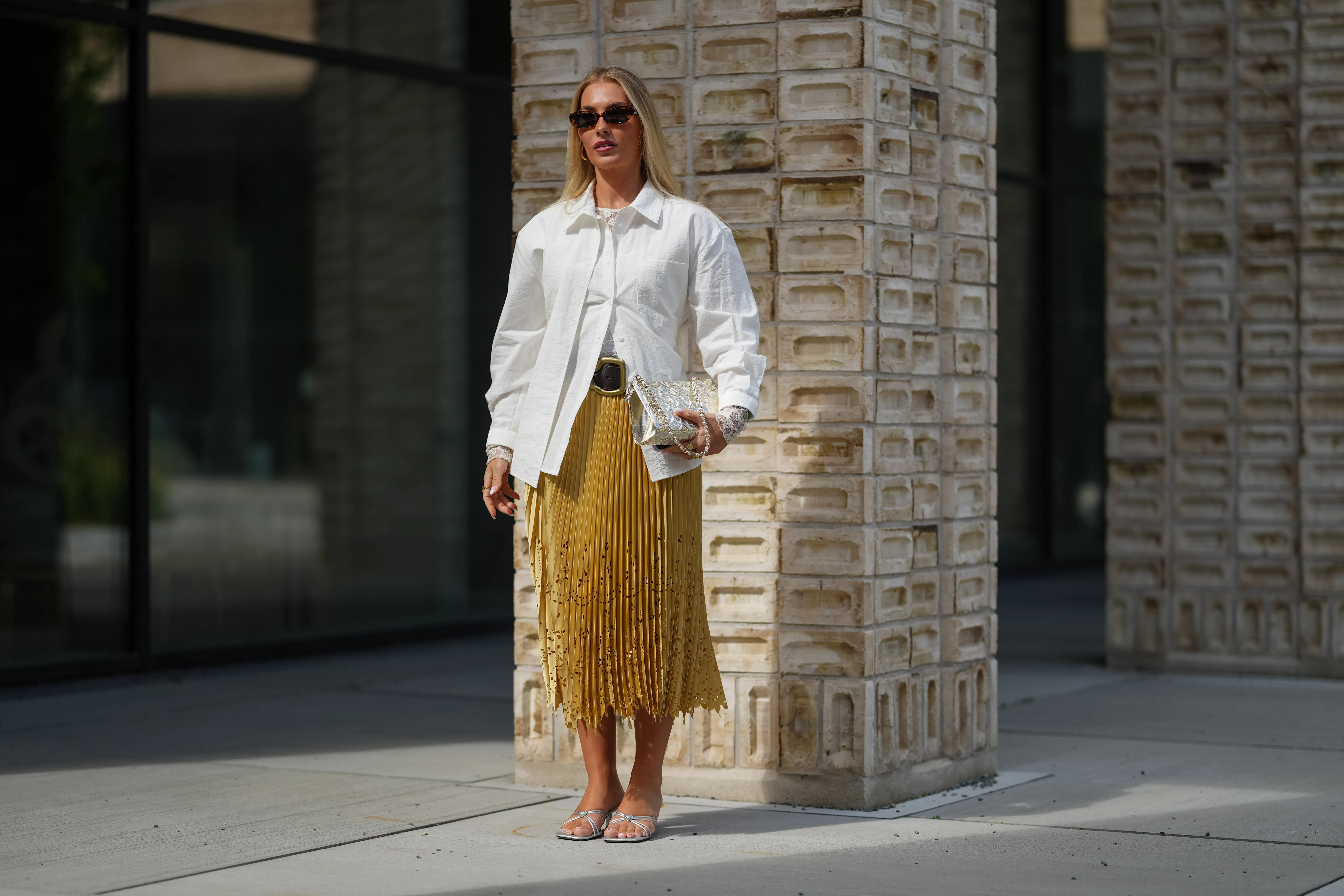 COPENHAGEN, DENMARK - AUGUST 06: A guest wears a white button-up shirt with a classic collar and long sleeves. The shirt is layered over a mustard yellow pleated skirt with intricate cut-out details near the hem. Sunglasses with dark lenses and a tortoiseshell frame are worn. A metallic clutch bag with a textured surface is held in one hand. The outfit is accessorized with a wide black belt featuring a prominent buckle. Footwear consists of silver strappy sandals with a low heel, outside Herskind, during Copenhagen Fashion Week, on August 06, 2025 in Copenhagen, Denmark (Photo by Edward Berthelot/Getty Images)