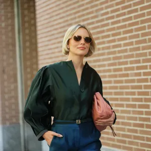 Violet Grace Atkinson wears a chin-length straight blonde bob with a center part, gold-framed brown-lens sunglasses, small gold stud earrings, a pink slouchy leather pouch bag, a dark green satin blouse with a slight stand collar, concealed front buttons and voluminous puff long sleeves, a thin black leather belt from Fendi with a gold-tone buckle and high-waisted teal satin wide-leg trousers, outside Fendi, during Milan Fashion Week - Womenswear Spring/Summer 2026, on September 24, 2025 in Milan, Italy
