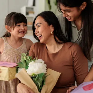 Heartwarming moment of two daughters giving their mother handmade cards, wrapped gifts, and a bouquet of flowers in a cozy living room. The family shares smiles and affection, celebrating Mother’s Day with love and appreciation. Perfect for concepts of motherhood, gratitude, family connection, and special occasions.