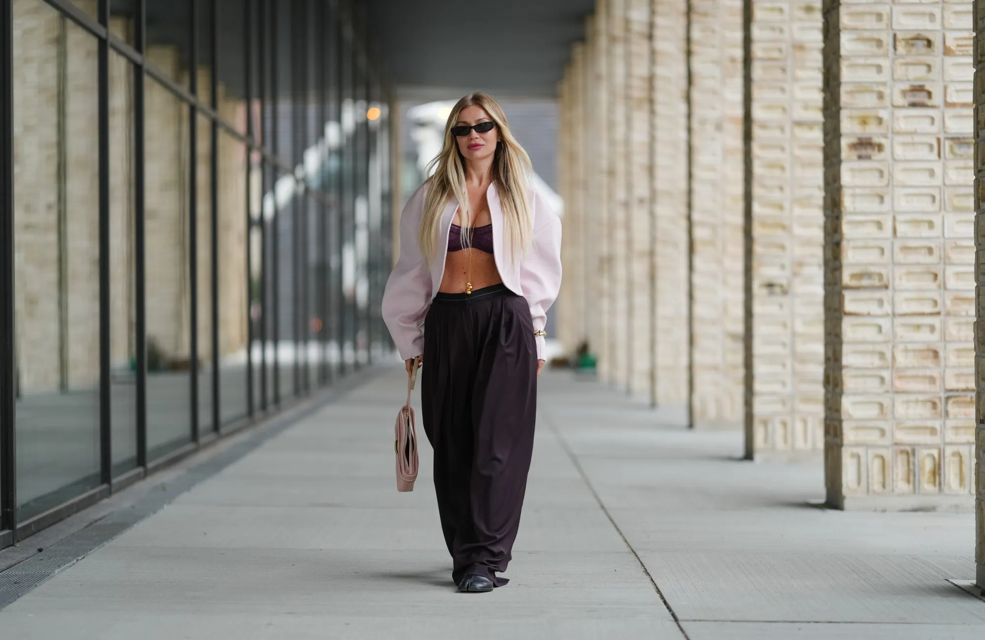 COPENHAGEN, DENMARK - AUGUST 07: A guest wears long, straight, center-parted blonde hair with darker roots, narrow black rectangular sunglasses, and pink lipstick; a pale pink cropped shrug-style jacket with rounded shoulders and voluminous sleeves is worn open over a burgundy ruched bandeau bralette, accented with a delicate gold body chain and a gold bracelet; high-waisted black wide-leg trousers in a fluid fabric are paired with black round-toe flat shoes; a blush pink structured leather top-handle bag with a flap and gold-tone hardware is carried in hand, walking, outside Birrot, during Copenhagen Fashion Week, on August 07, 2025 in Copenhagen, Denmark (Photo by Edward Berthelot/Getty Images)