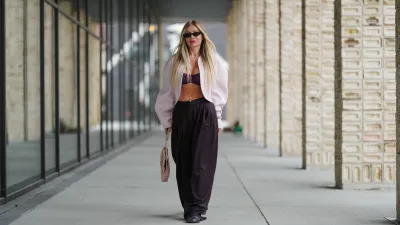 COPENHAGEN, DENMARK - AUGUST 07: A guest wears long, straight, center-parted blonde hair with darker roots, narrow black rectangular sunglasses, and pink lipstick; a pale pink cropped shrug-style jacket with rounded shoulders and voluminous sleeves is worn open over a burgundy ruched bandeau bralette, accented with a delicate gold body chain and a gold bracelet; high-waisted black wide-leg trousers in a fluid fabric are paired with black round-toe flat shoes; a blush pink structured leather top-handle bag with a flap and gold-tone hardware is carried in hand, walking, outside Birrot, during Copenhagen Fashion Week, on August 07, 2025 in Copenhagen, Denmark (Photo by Edward Berthelot/Getty Images)