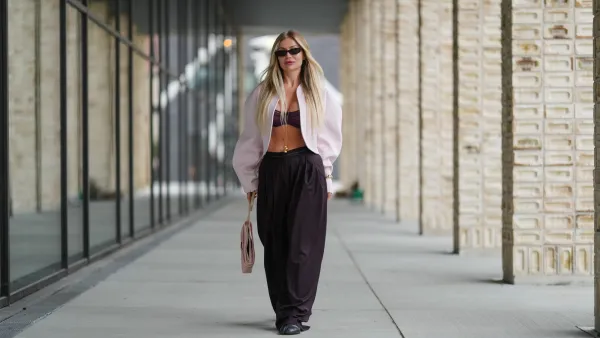 COPENHAGEN, DENMARK - AUGUST 07: A guest wears long, straight, center-parted blonde hair with darker roots, narrow black rectangular sunglasses, and pink lipstick; a pale pink cropped shrug-style jacket with rounded shoulders and voluminous sleeves is worn open over a burgundy ruched bandeau bralette, accented with a delicate gold body chain and a gold bracelet; high-waisted black wide-leg trousers in a fluid fabric are paired with black round-toe flat shoes; a blush pink structured leather top-handle bag with a flap and gold-tone hardware is carried in hand, walking, outside Birrot, during Copenhagen Fashion Week, on August 07, 2025 in Copenhagen, Denmark (Photo by Edward Berthelot/Getty Images)