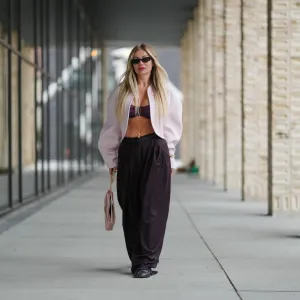 COPENHAGEN, DENMARK - AUGUST 07: A guest wears long, straight, center-parted blonde hair with darker roots, narrow black rectangular sunglasses, and pink lipstick; a pale pink cropped shrug-style jacket with rounded shoulders and voluminous sleeves is worn open over a burgundy ruched bandeau bralette, accented with a delicate gold body chain and a gold bracelet; high-waisted black wide-leg trousers in a fluid fabric are paired with black round-toe flat shoes; a blush pink structured leather top-handle bag with a flap and gold-tone hardware is carried in hand, walking, outside Birrot, during Copenhagen Fashion Week, on August 07, 2025 in Copenhagen, Denmark (Photo by Edward Berthelot/Getty Images)