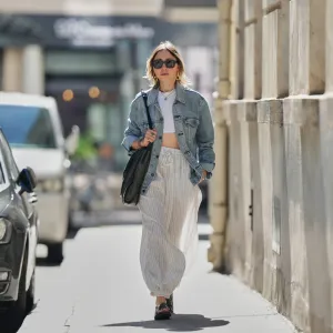 PARIS, FRANCE - SEPTEMBER 06: Emy Venturini wears straight light brown hair with a middle part, black vintage sunglasses, gold hoop earrings, layered necklaces, multiple bracelets, a large black vintage leather shoulder bag, a light blue denim jacket by Levi’s, a white cropped top, wide white striped drawstring pants by Zara Studio, and black leather cross-strap sandals by Prada, during a street style fashion photo session, on September 06, 2025 in Paris, France. (Photo by Edward Berthelot/Getty Images)