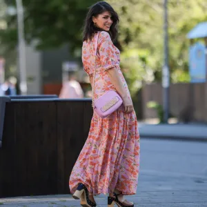 Diane Batoukina wears a white silk midi dress with blue and navy graphic stripe print, short sleeves, and a pointed collar by Sandro. A matching Sandro silk scarf is tied loosely around the neckline. A white mini flap bag with a chain strap and gold hardware by Chanel is worn crossbody. Footwear consists of white open-toe heeled sandals by Aeyde. Dior square sunglasses with dark gradient lenses complete the look. Long, straight brown hair is styled with a center part. Lips are subtly tinted and skin appears smooth and even. Full body shot, during a street style fashion photo session, on April 28, 2025 in Paris, France.