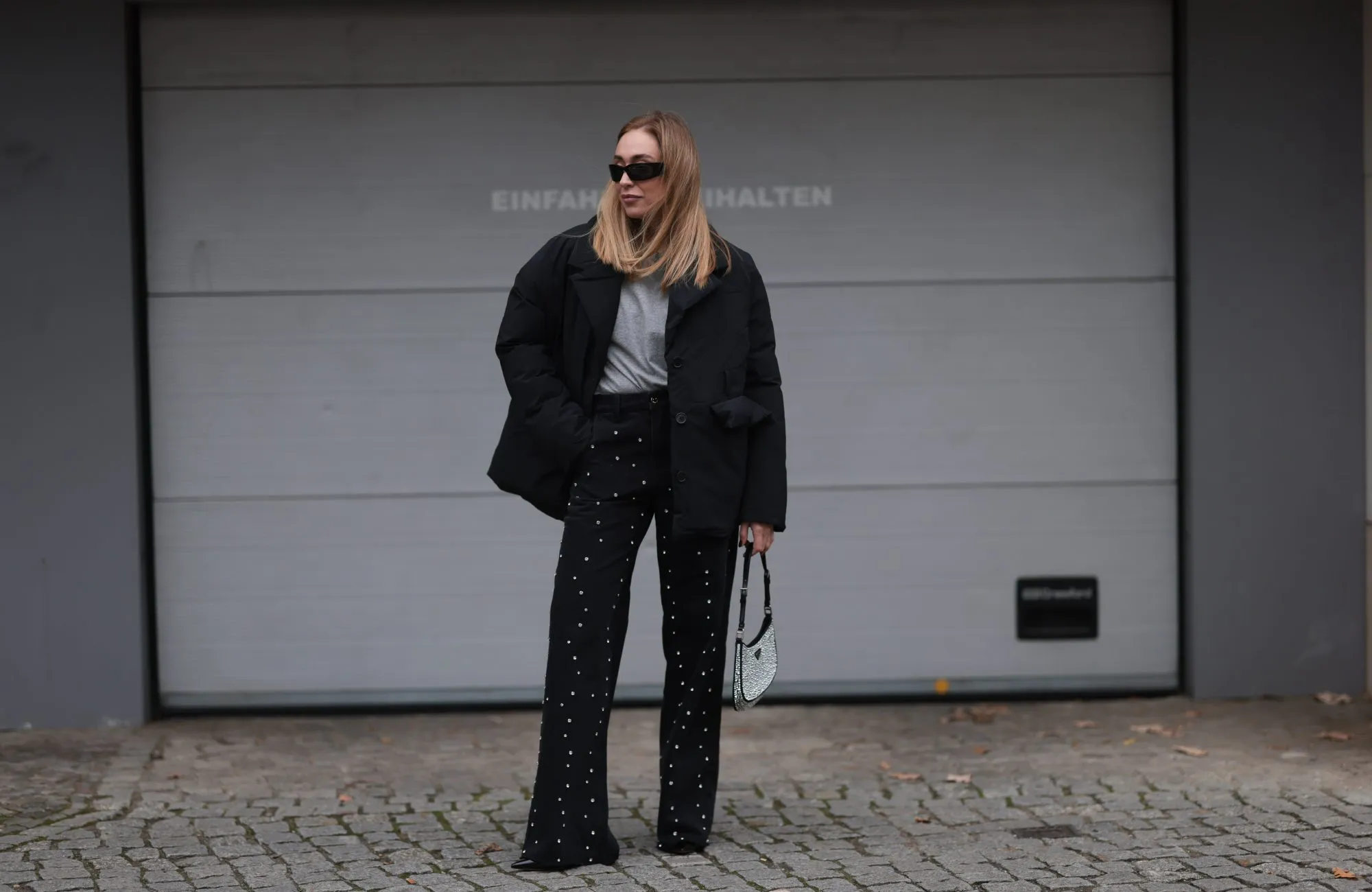 BERLIN, GERMANY - NOVEMBER 15: Sonia Lyson is seen wearing black rectangular sunglasses featuring wide temples with triangular logo embellishments from Prada; silver diamond-studded hoop earrings; a padded black jacket with notch lapels from Marina Hoermanseder x About You; underneath a grey t-shirt from Sézane; wide-legged straight black jeans with rhinestones pattern also from Marina Hoermanseder x About You; a crystal-embellished black leather “Cleo“ bag from Prada; and pointed black leather heels from Jimmy Choo on November 15, 2024 in Berlin, Germany. (Photo by Jeremy Moeller/Getty Images)