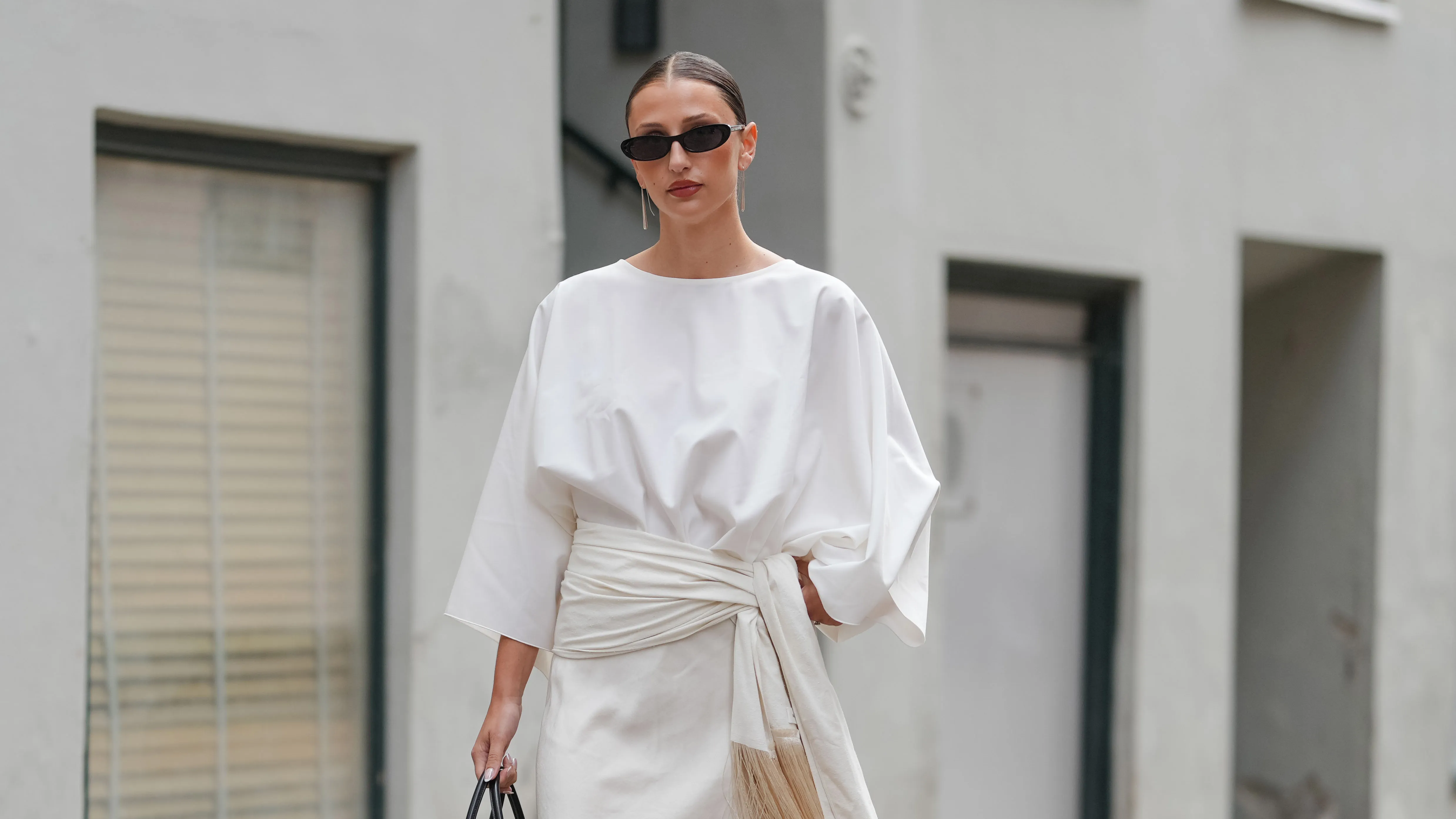 COPENHAGEN, DENMARK - AUGUST 04: A guest wears a white blouse with three-quarter length sleeves and a round neckline. The outfit includes a white skirt with a draped detail and fringe accents. A black shiny leather Birkin handbag from Hermes with a structured shape is held. Black sunglasses with a rectangular frame are worn. Black patent leather heels complete the ensemble. Hair is styled in a sleek, pulled-back manner, outside Freya Dalsjo, during Copenhagen Fashion Week, on August 04, 2025 in Copenhagen, Denmark (Photo by Edward Berthelot/Getty Images)