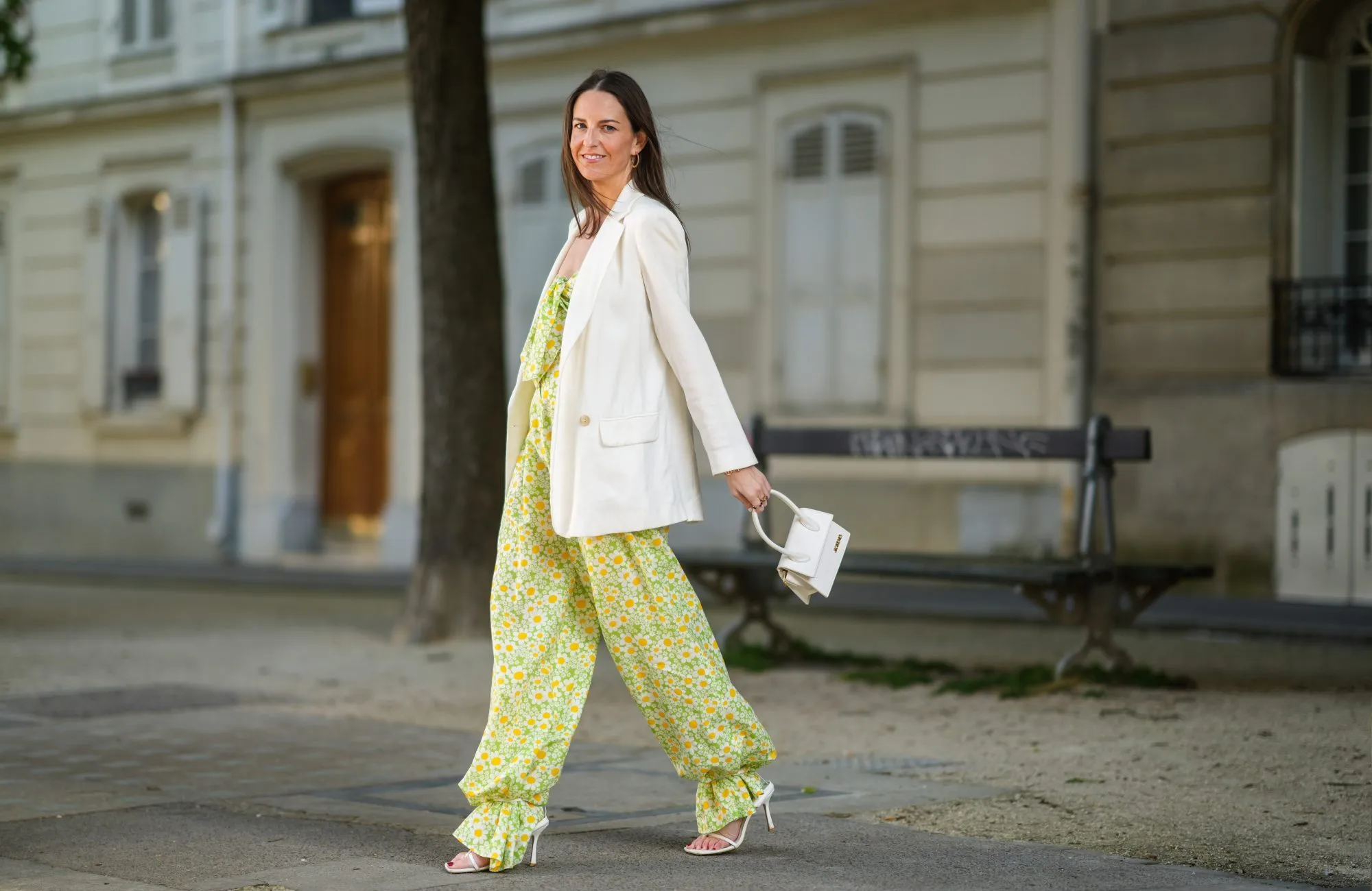 PARIS, FRANCE - APRIL 22: Alba Garavito Torre wears gold earrings, a green with white daisy pattern Mirae jumpsuit with off shoulder knotted neck and puffy ankles, a white long blazer jacket, a gold and rhinestones bracelet, gold rings, a gold pink APM Monaco bracelet, a white shiny leather Le Chiquito medium Jacquemus handbag, white matte leather strappy Bottega Veneta sandals heels pumps shoes, on April 22, 2021 in Paris, France. (Photo by Edward Berthelot/Getty Images)