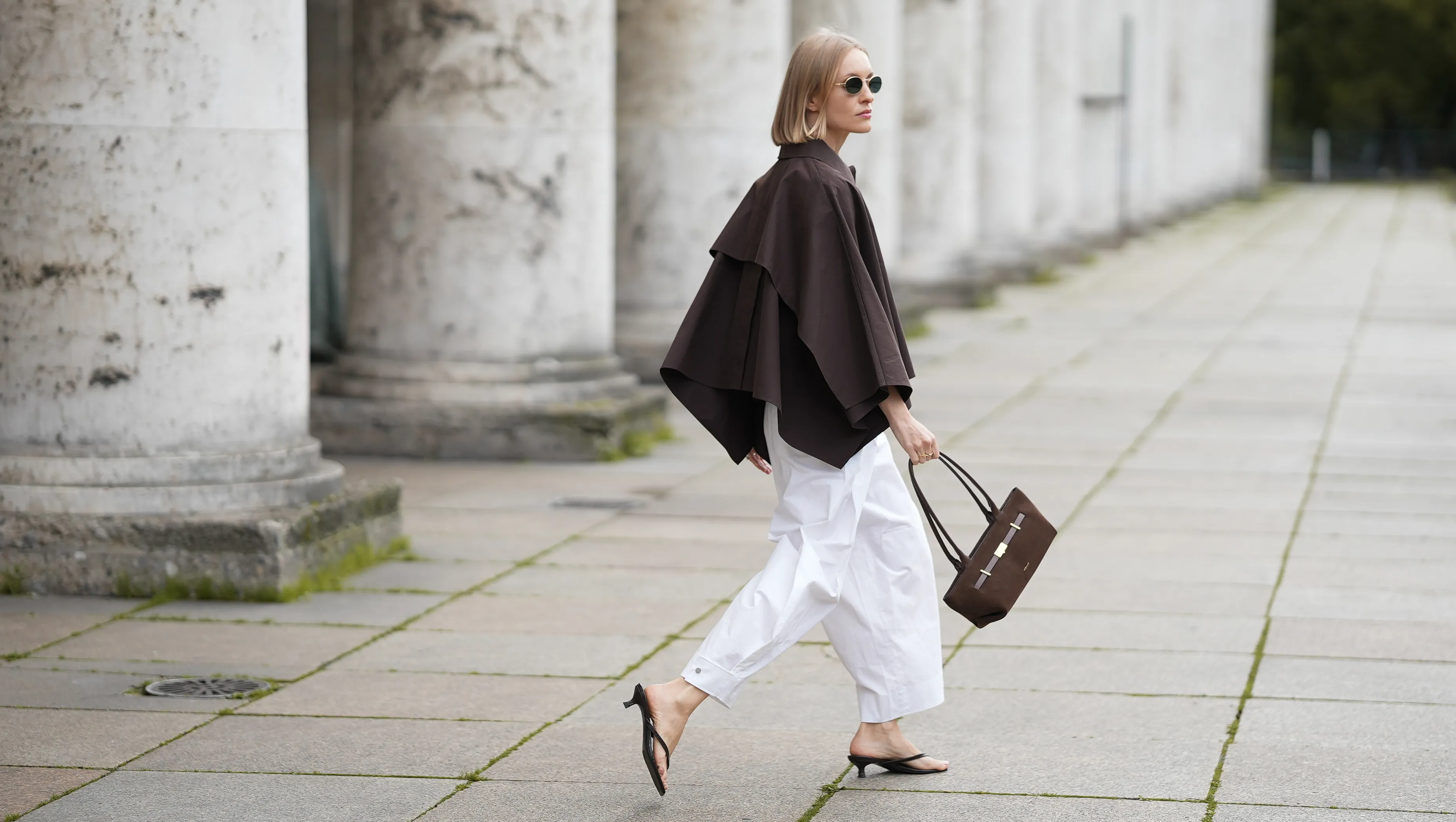 MUNICH, GERMANY - SEPTEMBER 2: Marlies Pia Pfeifhofer is seen wearing a voluminous dark brown cropped trench coat cape with wide draped sleeves from COS; white barrel trousers with ankle length and wide leg silhouette from COS; a pair of black flip-flop heeled sandals with square toes from H&M; a dark brown suede The New York shoulder bag with top handles, triangular fold design and golden hardware from DeMellier; round sunglasses with slim golden frames and dark-shaded lenses from Persol; her blonde bob hair is styled in a sleek, chin-length cut with a center parting on September 2, 2025 in Munich, Germany. (Photo by Moritz Scholz/Getty Images)